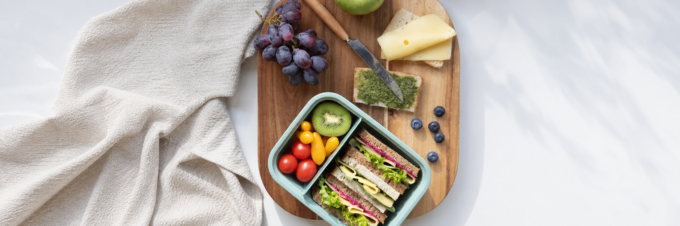 Mint green lunch box filled with sandwiches, fresh fruit and tomatoes on a wooden board.