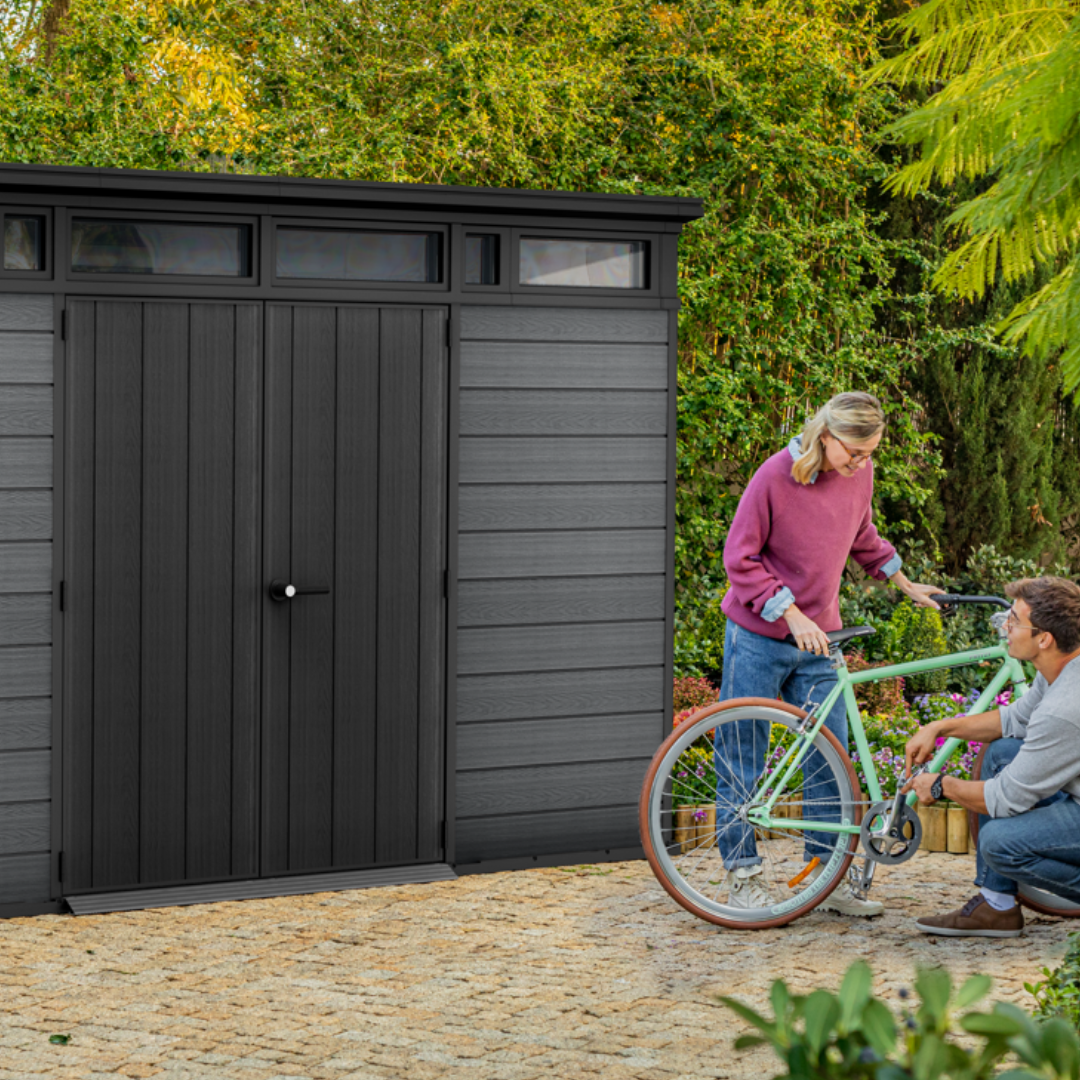 Front view of the Cortina 11x7ft garden shed with a family and bicycle beside it in a backyard setting