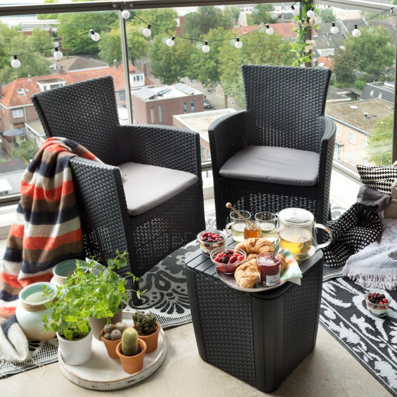 Iowa Balcony Set with two dark flat wicker armchairs and a matching storage side table arranged on a balcony with cushions and breakfast items