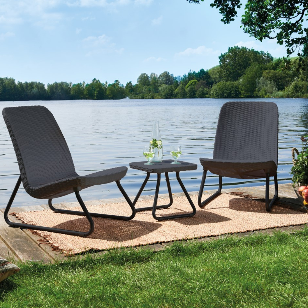 Rio Patio Set in graphite with two wicker-look chairs and a matching side table arranged beside a lakeside garden