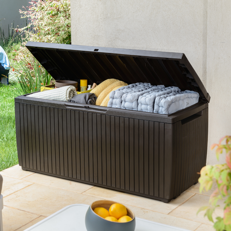 Springwood 305L Storage Box with open hinged lid revealing cushions and blankets in a dark brown slatted finish on a patio