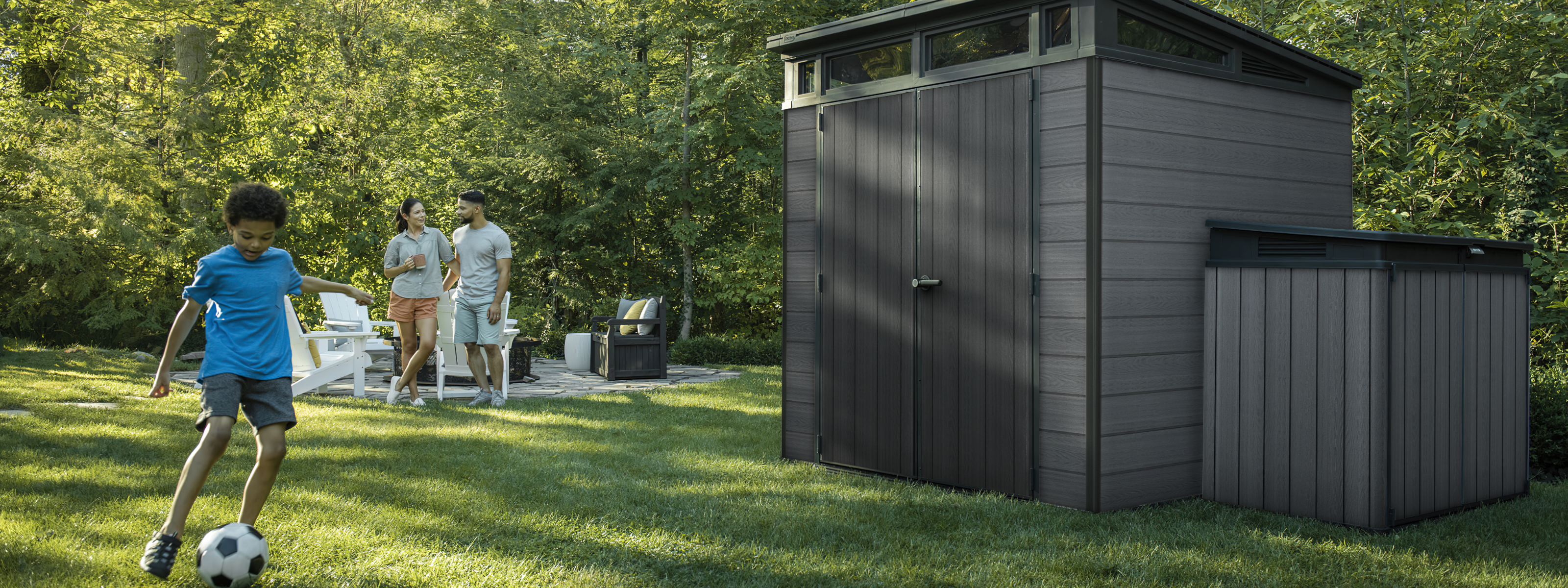 Children playing soccer on the lawn next to a modern grey Keter garden shed, with family relaxing in the background.