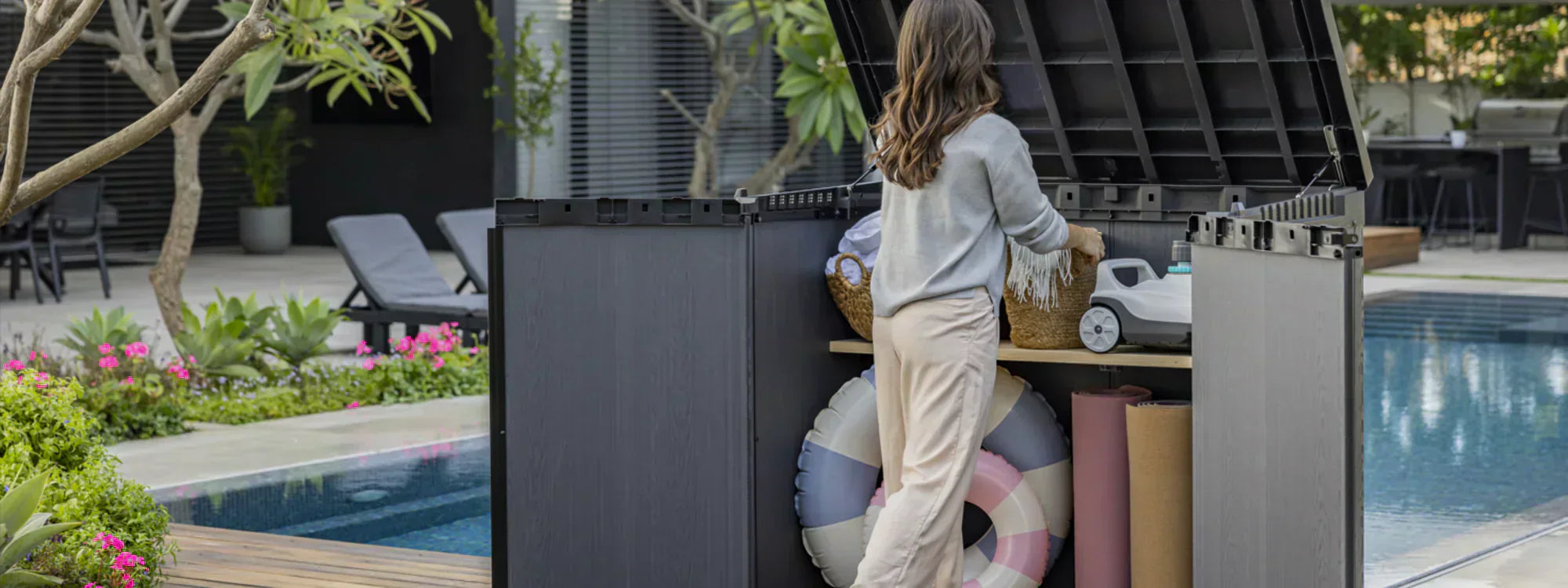Woman organising outdoor items in a large Keter storage box beside a pool and patio seating area.