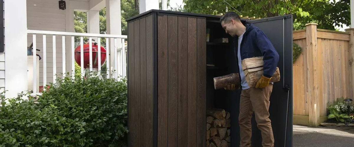 Man placing firewood into a durable storage shed beside a backyard patio and garden area.