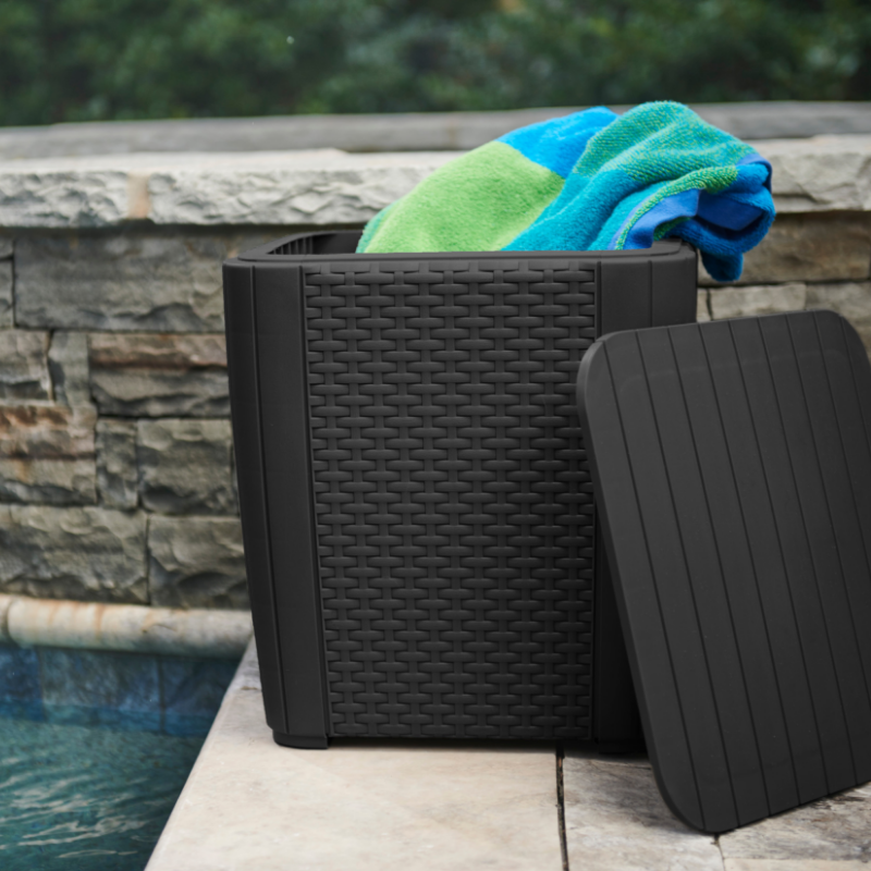 Black woven-effect storage box with a ribbed lid leaning against it beside a swimming pool and rolled towels inside